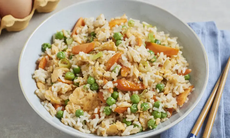 Schüssel mit gebratenem Eierreis wie beim Chinesen, garniert mit Erbsen und Karotten. Daneben liegen Stäbchen.