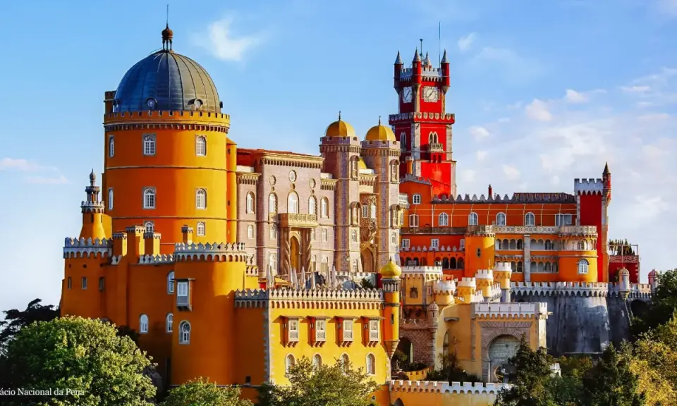 Buntes Sintra Pena Palace mit gelben und roten Türmen, einer blauen Kuppel und einem Uhrenturm unter blauem Himmel.