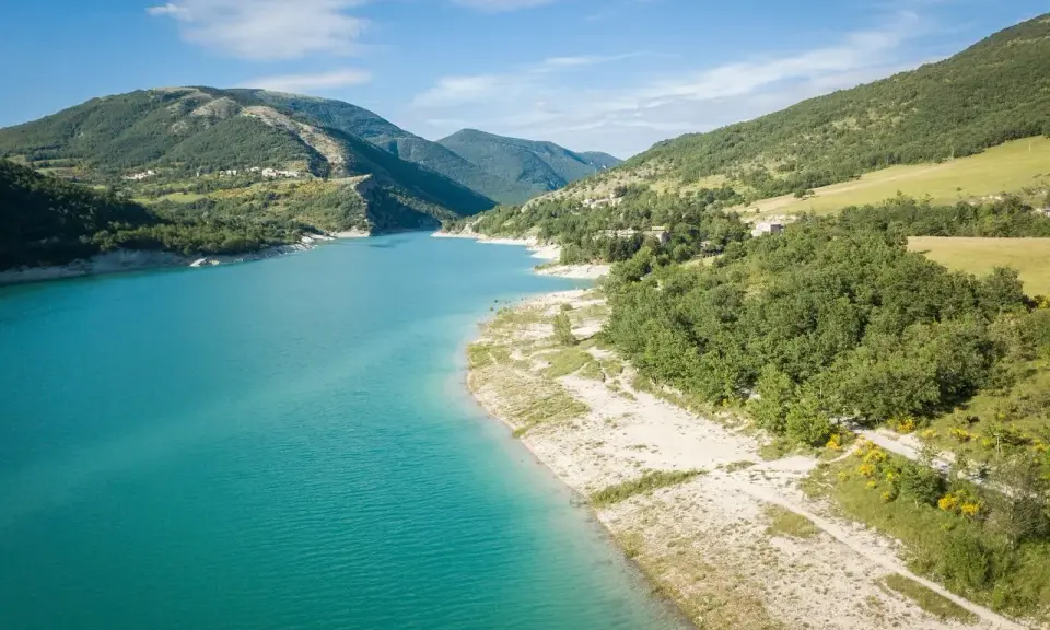 Splendido **lago di Fiastra** con acque turchesi, circondato da colline verdi e case sparse. Un paesaggio sereno e incantevole.