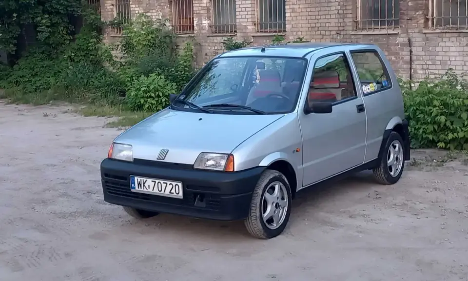 Srebrne cinquecento z czerwonymi akcentami we wnętrzu, zaparkowane na piaszczystym podwórku przed ceglanym budynkiem.