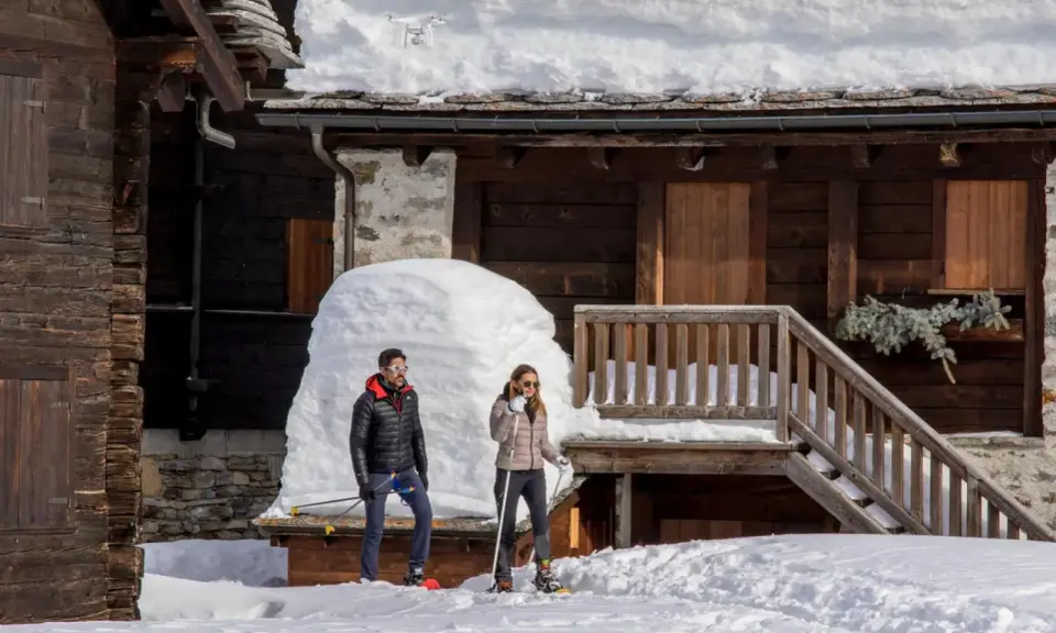 Coppia con racchette da neve a Riale Val Formazza, con un drone in volo sopra un tetto innevato.