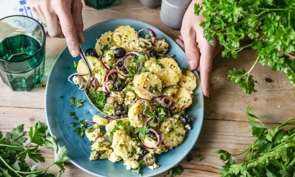 Ein köstlicher mediterraner Kartoffelsalat mit Oliven, roten Zwiebeln und Feta auf einem blauen Teller, garniert mit frischer Petersilie.
