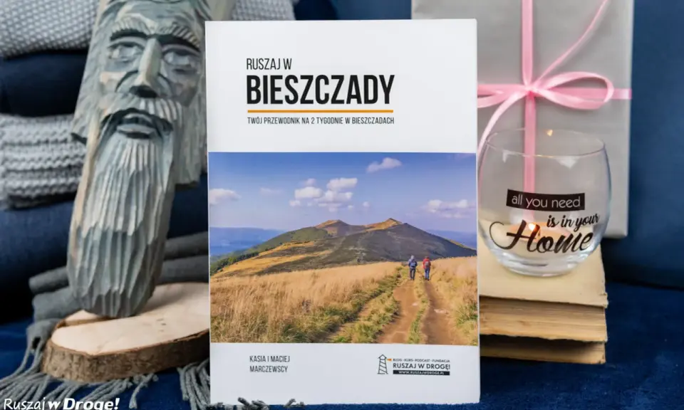 Przewodnik "Ruszaj w Bieszczady" z widokiem na góry i turystów. Obok rzeźba z drewna i świeca z napisem "all you need is in your Home".