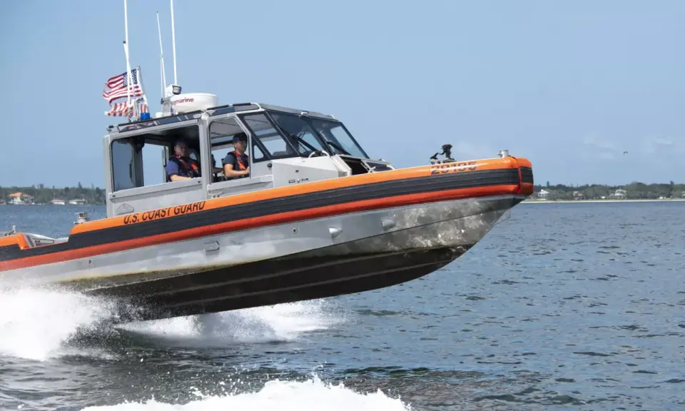 Un bateau du garde côte américain file sur l'eau, créant une gerbe d'écume. Deux membres d'équipage sont visibles à l'intérieur.