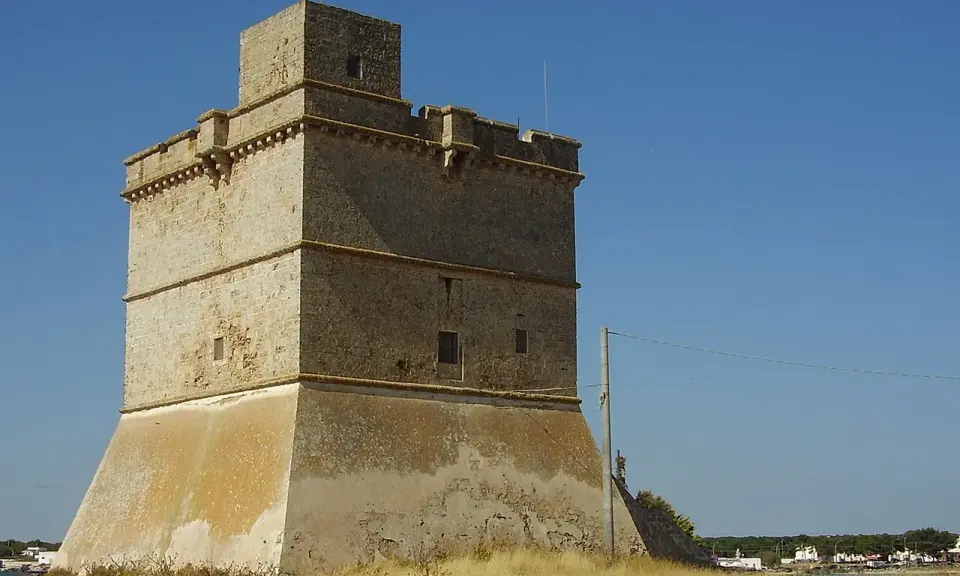 Torre costiera a Sant Isidoro, Salento. Antica sentinella di pietra contro il cielo azzurro, con il mare e il borgo sullo sfondo.