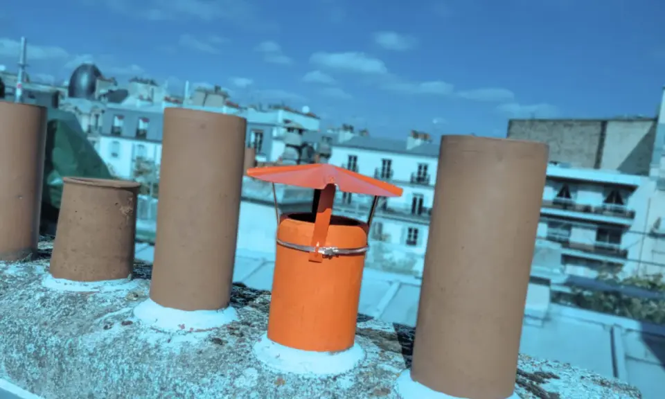 Sur un toit parisien, un chapeau chinois orange protège une cheminée, entouré d'autres conduits sous un ciel bleu.