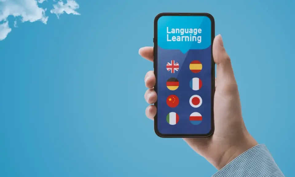 Dłoń trzyma smartfon z aplikacją do nauki języków. Wybór między Busuu czy Duolingo? Na ekranie flagi: UK, Hiszpanii, Niemiec, Francji, Chin, Japonii, Włoch, Polski.