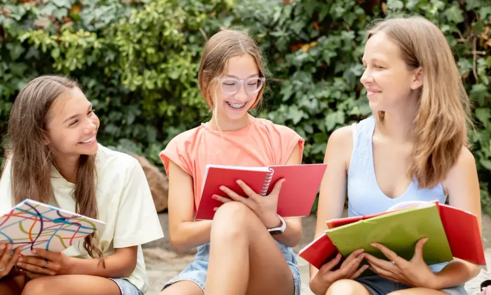 Tre ragazze sorridono studiando insieme, circondate dal verde. Usano quaderni colorati, ottime risorse didattiche Pearson per imparare all'aperto.