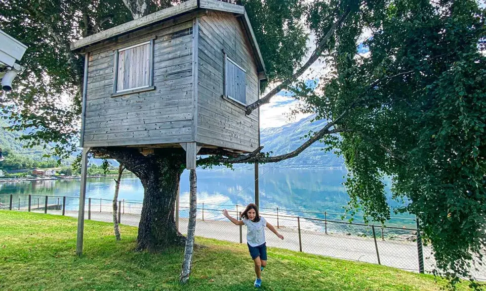 Bambina corre verso una casa sull'albero in legno, con un lago e montagne sullo sfondo.