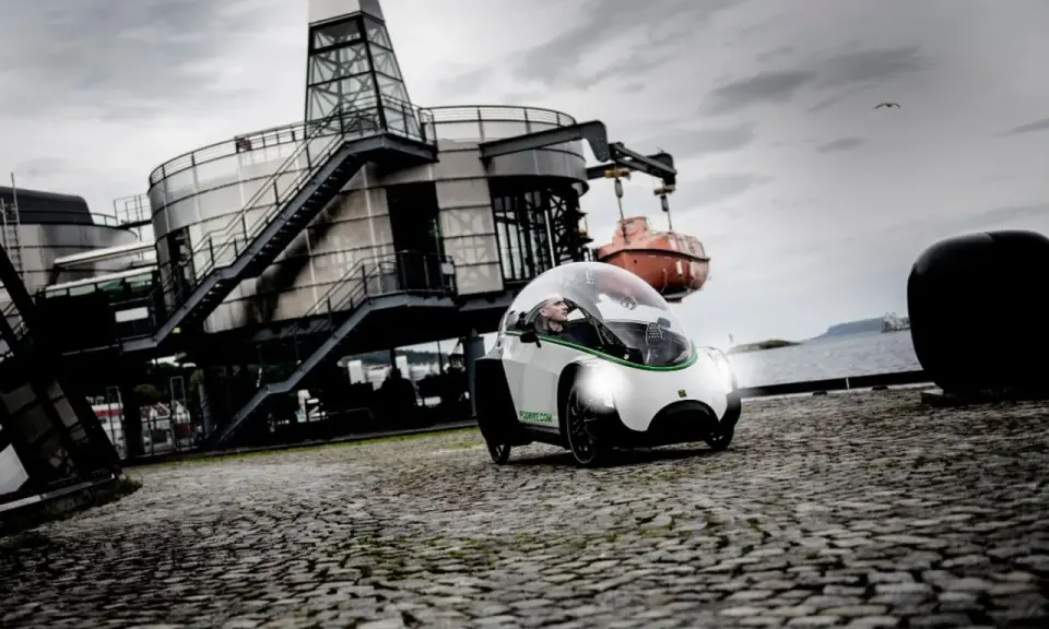 Ein Podbike Frikar fährt auf Kopfsteinpflaster vor einem modernen Gebäude am Wasser.