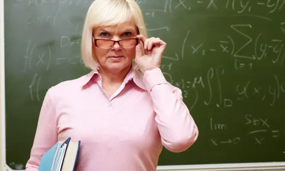 Nauczycielka matematyki poprawia okulary, trzymając książki. Na tablicy widać wzory. Dziś jest 14 października.