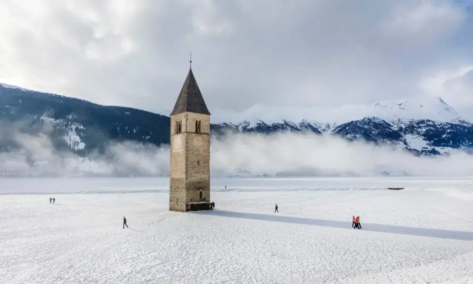 Torre sommersa nel lago di Resia, circondata da neve e montagne nebbiose. Alcune persone passeggiano sul ghiaccio.