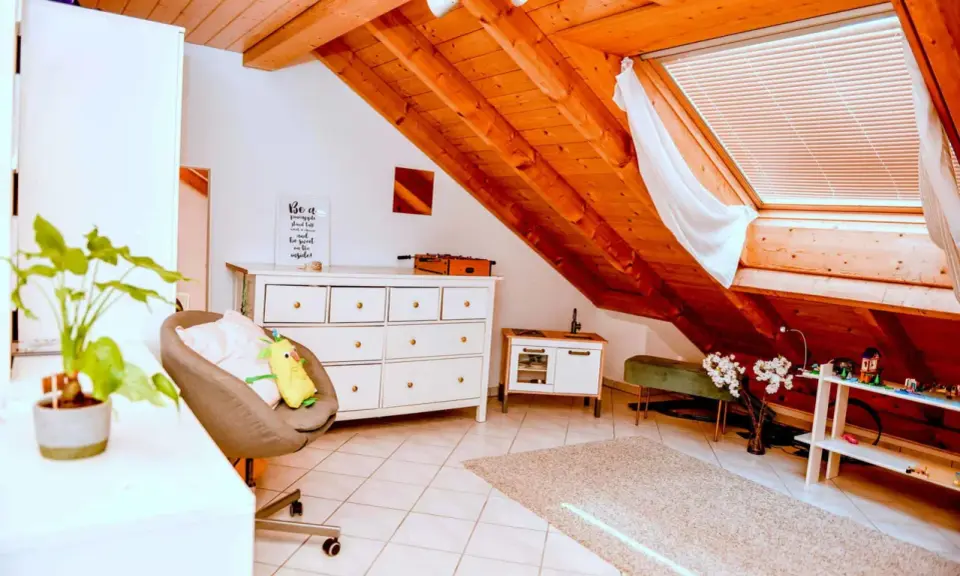 Acogedor despacho en buhardilla con vigas de madera, cómoda blanca, sillón y alfombra.