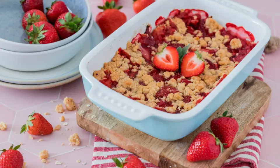 Ein frisch gebackener Erdbeer Crumble in einer hellblauen Auflaufform, garniert mit Erdbeerhälften. Daneben eine Schüssel voller Erdbeeren.
