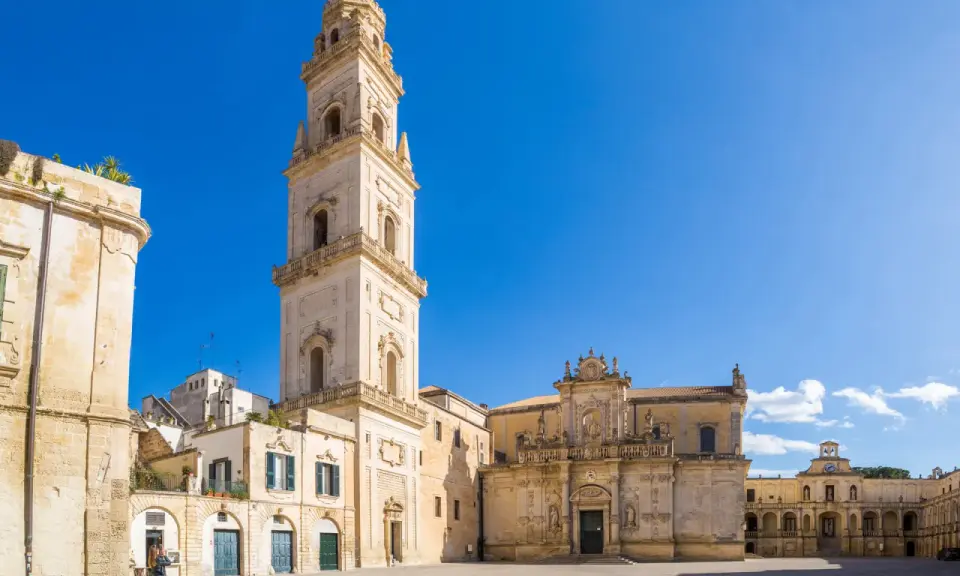 La piazza Carmelo Bene a Lecce, con la maestosa Basilica di Santa Croce e il campanile che svetta sotto un cielo azzurro.
