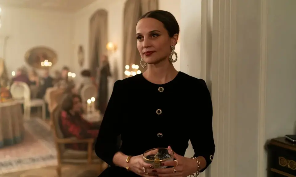 Alicia Vikander i en elegant svart klänning med stora örhängen, håller ett glas. Scenen utspelar sig på en tillställning, kanske från en av hennes filmer.