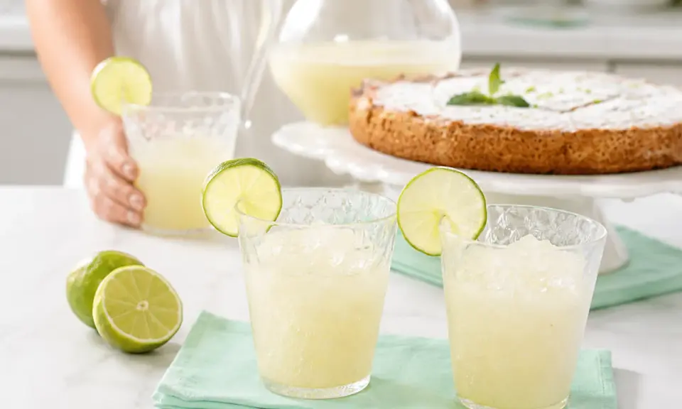 Refrescante granizado de limón Thermomix, perfecto para un día caluroso. Servido en vasos con rodajas de lima y acompañado de un delicioso pastel.