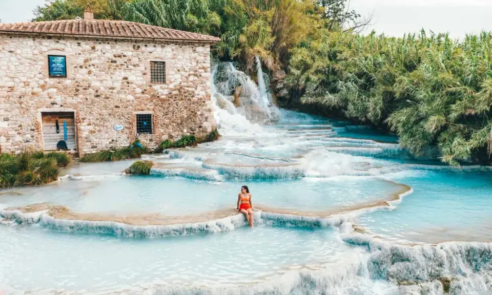 Cascate naturali con acqua turchese, una donna seduta su una roccia. Un antico mulino in pietra sullo sfondo. Tra le terme più belle d'Italia.