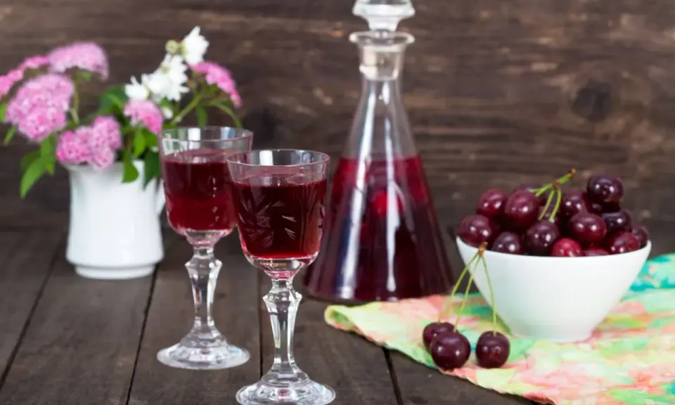 Dwa kieliszki z rubinową nalewką z czereśni na bimbrze, obok miseczka świeżych owoców i bukiecik kwiatów.