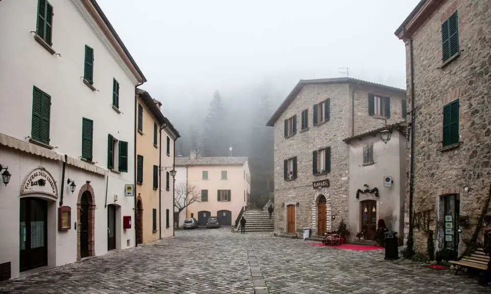 Piazza di San Leo avvolta nella nebbia, con edifici in pietra e intonaco, un bar e una farmacia.