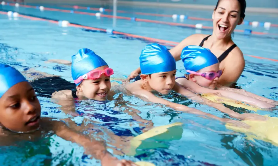 Fröhliche Kinder mit blauen Badekappen üben das schwimmen in der Grundschule. Eine Lehrerin hilft ihnen im Wasser.