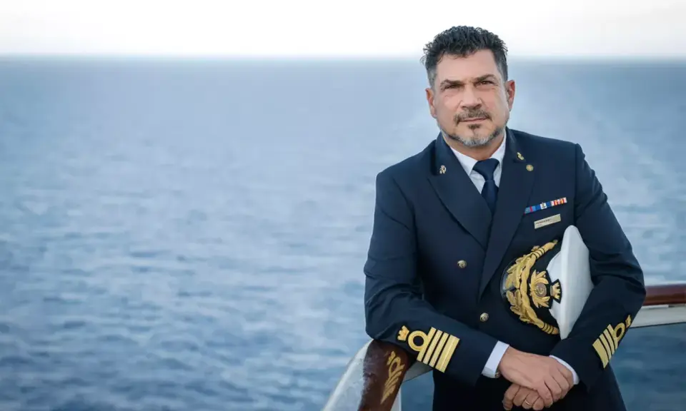 Il comandante MSC Splendida, con la sua uniforme blu scuro e il cappello dorato, posa con il mare calmo sullo sfondo.