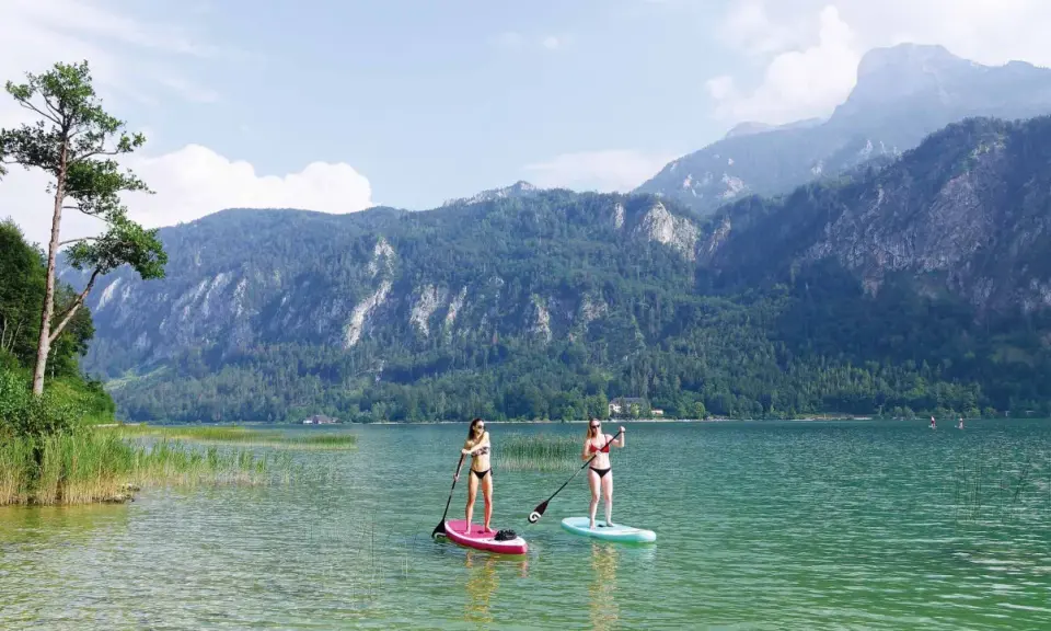 Zwei Frauen genießen Stand-Up-Paddling auf einem ruhigen See vor einer Bergkulisse. Perfekte sup spots für einen entspannten Tag.