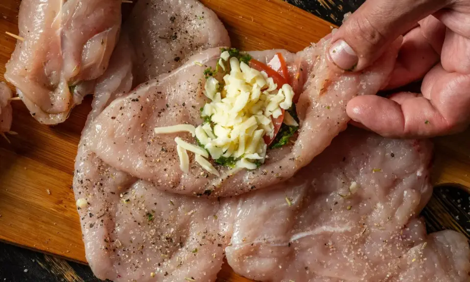 Przygotowanie roladki z kurczaka: ręka dodaje ser i pomidora do nadzienia na surowym mięsie.