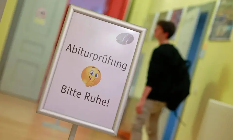 Schild mit der Bezeichnung "Abiturprüfung. Bitte Ruhe!" und einem Emoji, das zum Schweigen auffordert. Ein Schüler geht im Hintergrund vorbei.