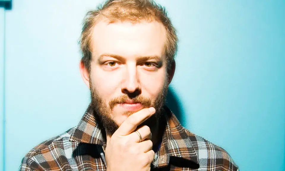 Bon Iver Sverige: en man med skägg och flanellskjorta funderar mot en turkos bakgrund.