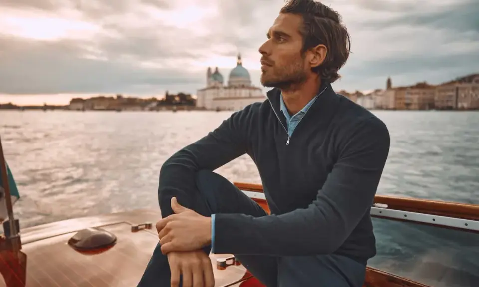 A man in mens dressy casual attire, a navy quarter-zip sweater over a blue collared shirt, sits on a boat with Venice in the background.