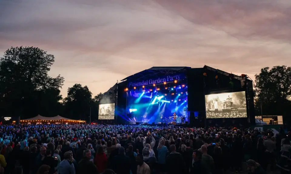Stor publik vid Rosendal Garden Party. En scen med blå ljus och artister, omgiven av träd och en vacker kvällshimmel.