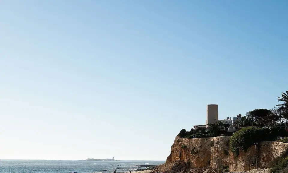 Sonniger Tag am **Playa La Barrosa**. Menschen genießen den Strand, während im Hintergrund eine Festung am Meer thront.