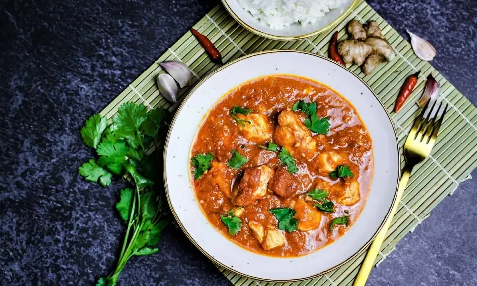 Ein Teller mit Hähnchen-Curry in cremiger **masala sauce**, garniert mit Koriander. Daneben Reis, Ingwer, Knoblauch und Chilis.