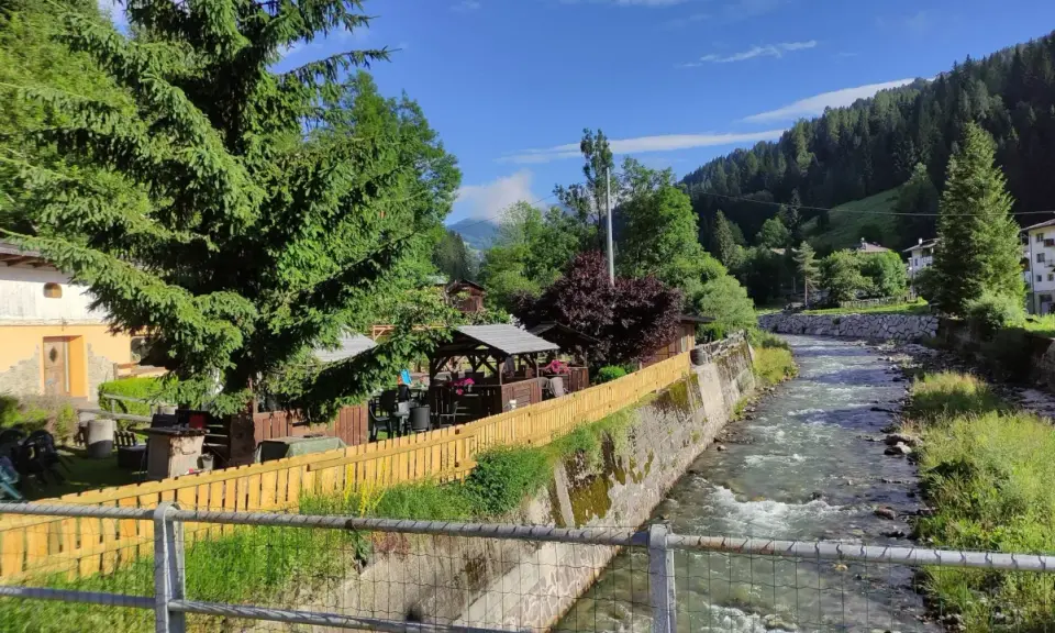 Un ruscello scorre a Santo Stefano di Cadore, fiancheggiato da case, alberi e un'area con gazebo e tavoli all'aperto.