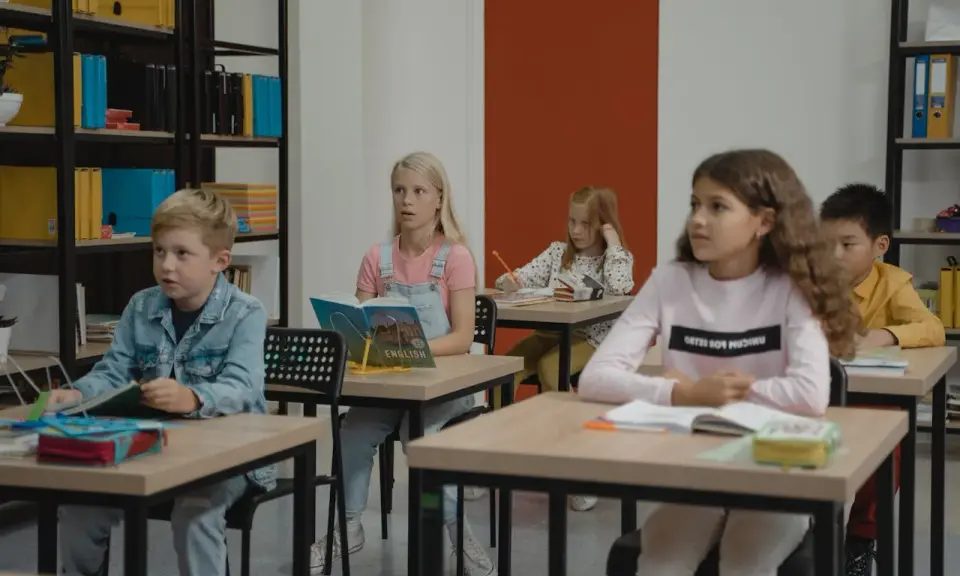 Dzieci siedzą w klasie, skupione na lekcji. Chłopiec w dżinsowej kurtce, dziewczynka z książką po angielsku, a z tyłu inne dzieci. Wszyscy mieli nadzieję, że lekcja szybko się skończy.