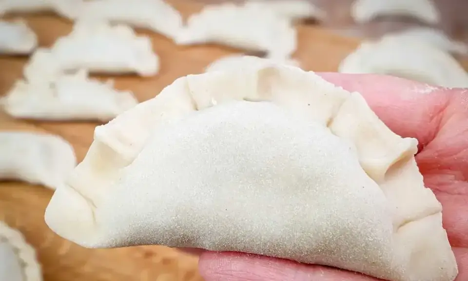 Dłoń trzyma surowe pierogi z serem, gotowe do gotowania. Na drewnianej desce widać więcej pierogów.