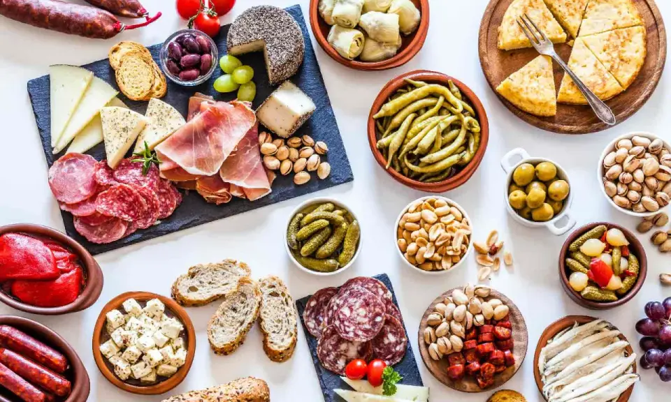 Una mesa llena de aperitivos baratos para mucha gente: embutidos, quesos, aceitunas, frutos secos, pimientos, pepinillos y pan.