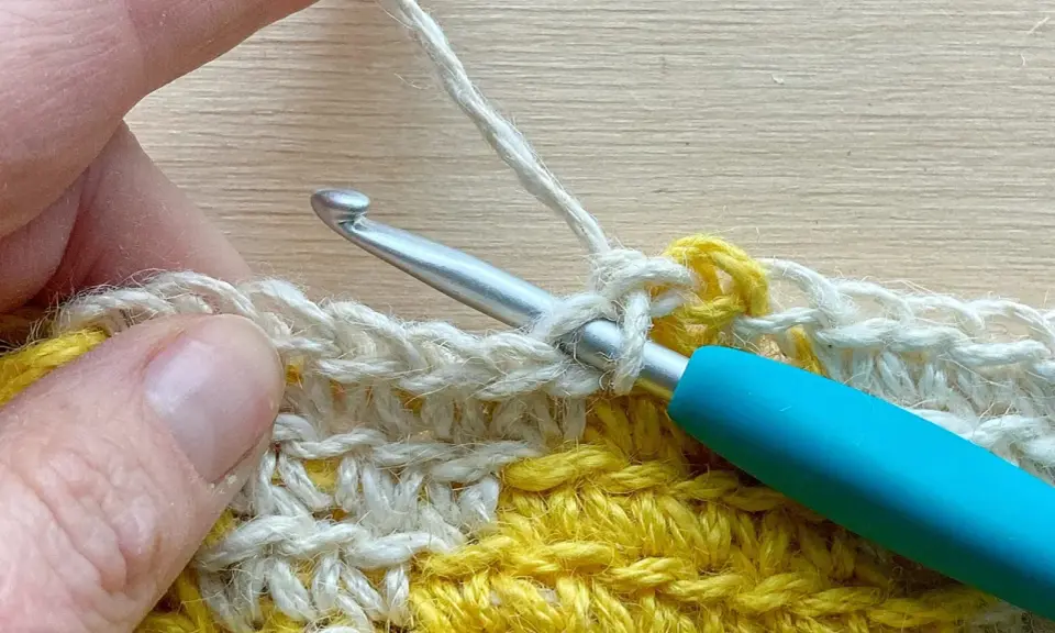 Mano tejiendo con ganchillo, creando un punto bajo con hilo blanco sobre una tela amarilla y blanca.
