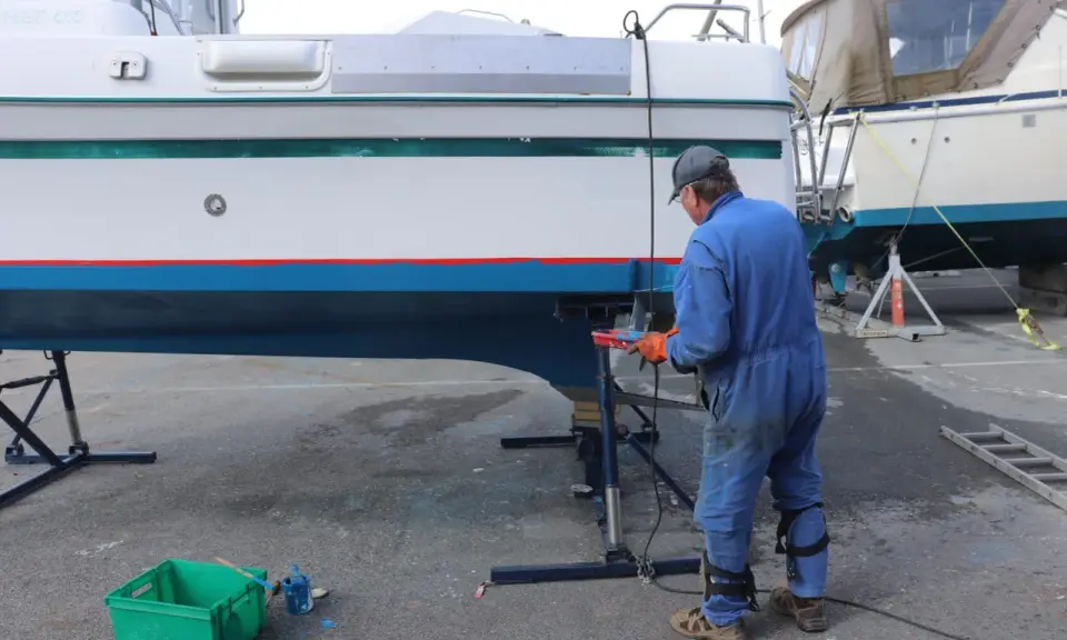 Un homme en combinaison bleue travaille sur la coque d'un bateau pour enlever l'antifouling. Des outils et des pots de peinture sont à proximité.
