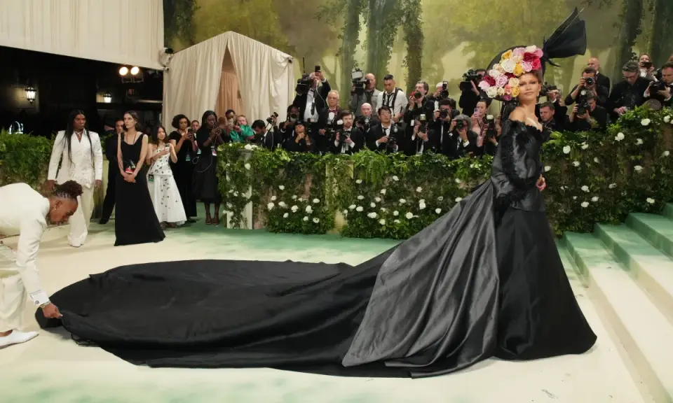 Una mujer con un vestido negro y un tocado floral posa para las cámaras en la alfombra verde, mientras un hombre ajusta la cola de su vestido, todo bajo el tema de la Met Gala.