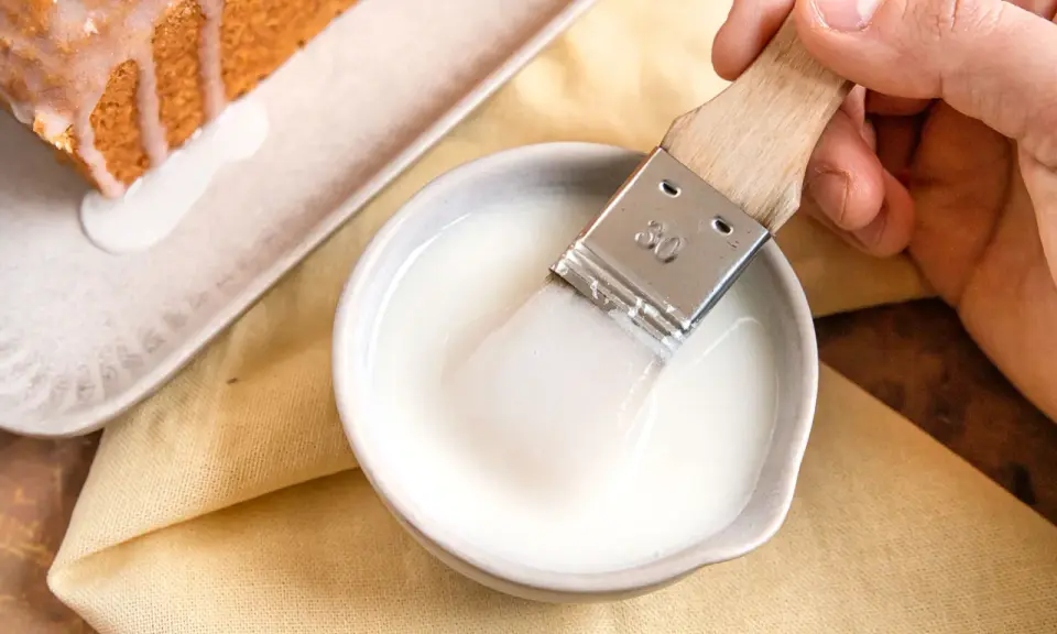Ein Pinsel taucht in eine Schüssel mit zuckerguss, bereit, einen warmen oder kalten Kuchen zu verfeinern.
