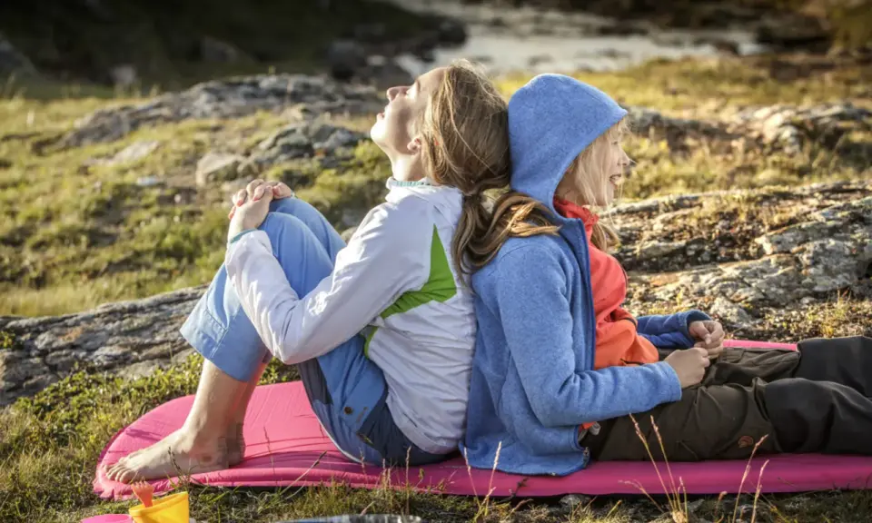 Två personer, en vuxen och ett barn, sitter rygg mot rygg på en liggunderlag i naturen. En härlig stund att vandra med barn.