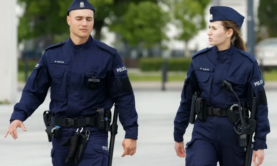 Dwóch policjantów w mundurach idzie, jeden z nich ma na pasie rękawiczki i pałkę.