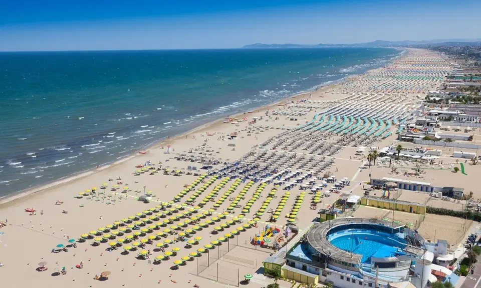 La riviera di Rimini in pieno sole, con un mare azzurro e una spiaggia affollatissima di ombrelloni colorati e bagnanti.