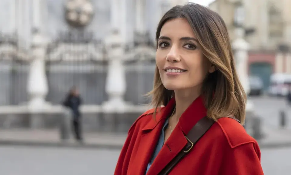 Donna con cappotto rosso sorride, con uno sfondo sfocato di edifici e una persona. Forse sta pensando ai **mina settembre libri** da leggere.