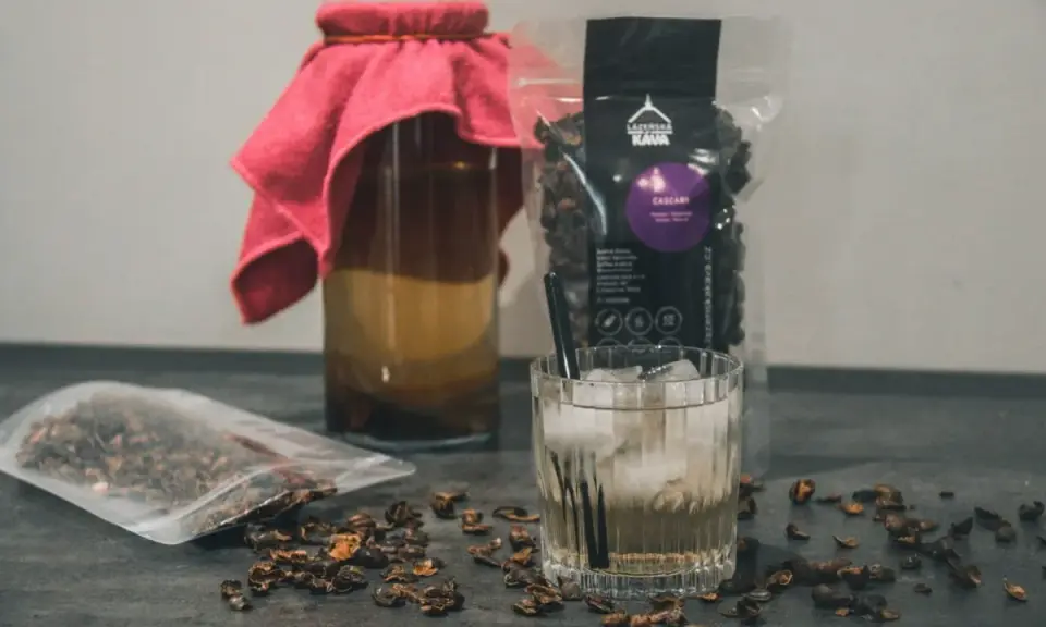 Napój z cascary, czyli co to jest kombucha z kawy, w szklance z lodem i słomką. Obok słoik z napojem i paczka z cascara.