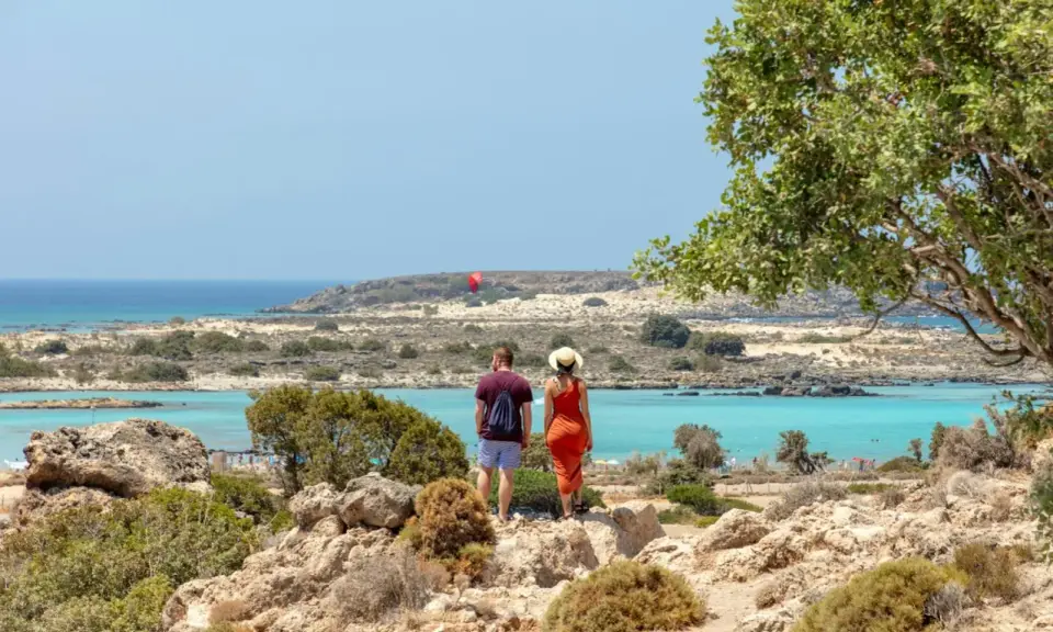 Ett par njuter av utsikten över Elafonissi, Kreta, med dess turkosa vatten och steniga landskap. En röd fallskärm syns i fjärran.
