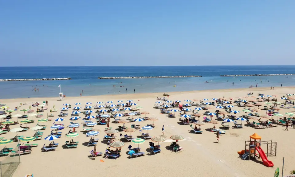 Spiaggia libera Viserba affollata di ombrelloni e lettini sotto un cielo azzurro. Bambini giocano nell'area attrezzata.