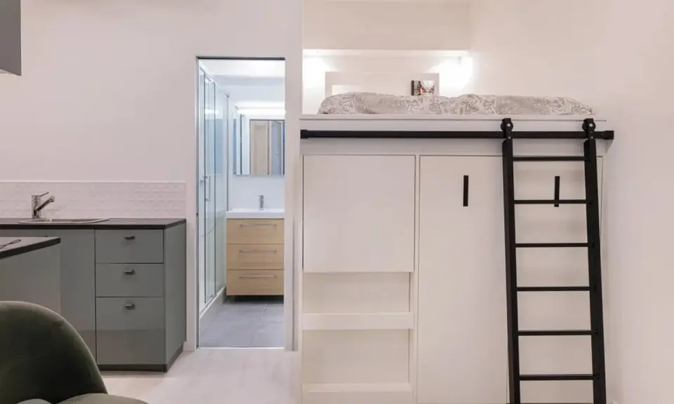 Un appartement fonctionnel avec lit mezzanine, échelle noire, cuisine grise et salle de bain visible.